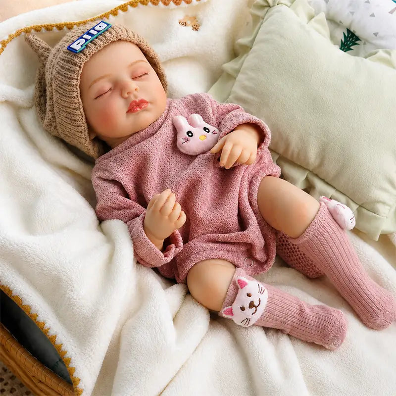 Sleeping reborn baby doll wearing a pink knitted romper, cat-ear hat, and pink cat socks, lying on a soft white blanket with a small bunny toy.