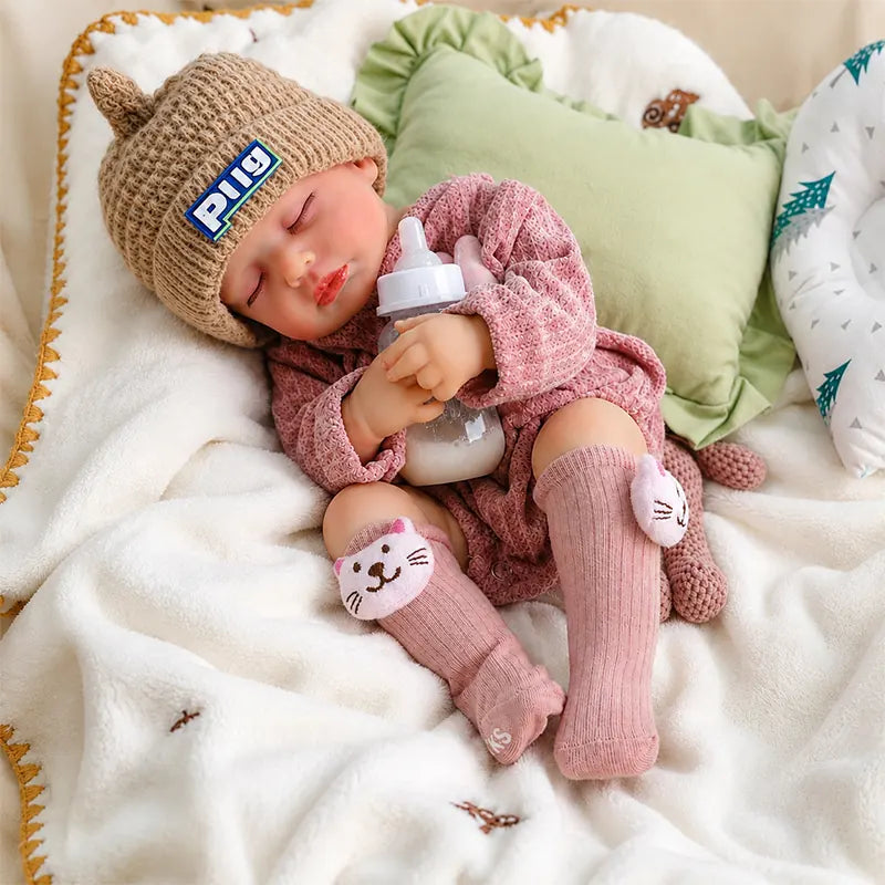 Sleeping reborn baby doll wearing a pink knitted outfit, cat-ear hat, and pink cat socks, cuddling a baby bottle on a soft white blanket.