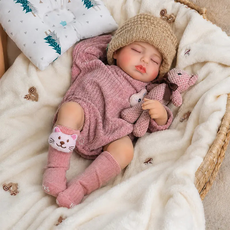 Sleeping reborn baby doll wearing a beige knitted hat, pink romper, and cat socks, cuddling a small knitted teddy bear on a soft white blanket.