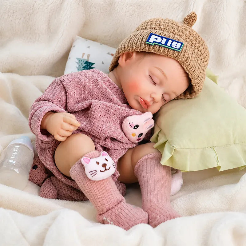 Sleeping reborn baby doll wearing a beige knitted hat, pink romper, and cat socks, resting beside a green pillow with a baby bottle nearby.