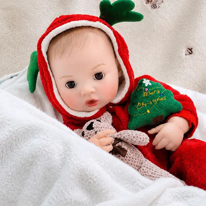Christmas interactive baby doll dressed in a red reindeer romper, holding a knitted bunny while sitting on soft white bedding.