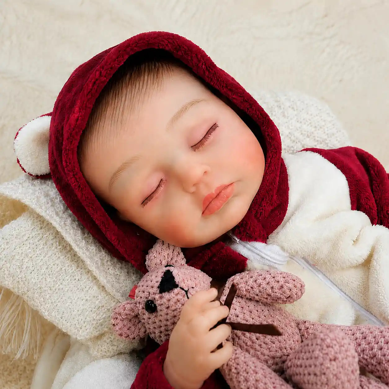 Sleeping reborn baby doll in red hooded outfit with bear ears, holding a pink crochet bunny, wrapped in cream knit blanket