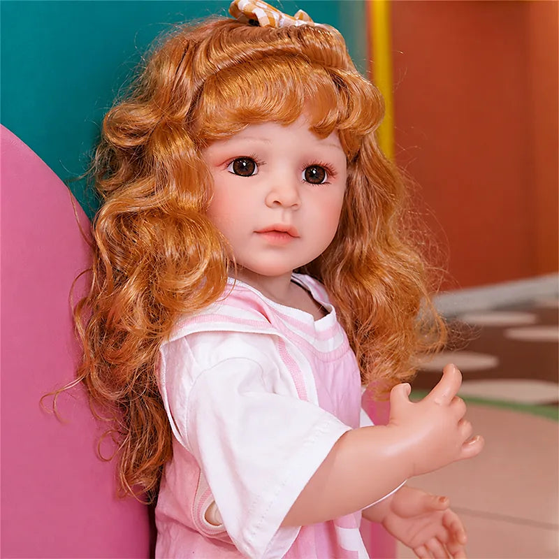 Adorable reborn toddler doll with bright eyes, voluminous auburn curls, a gingham bow, pink overalls, and white sneakers.
