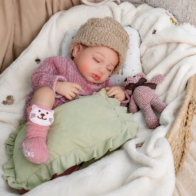 Sleeping reborn baby doll wearing a beige knitted hat, pink outfit, and cat socks, resting with a green pillow and a small knitted teddy bear on a soft white blanket.