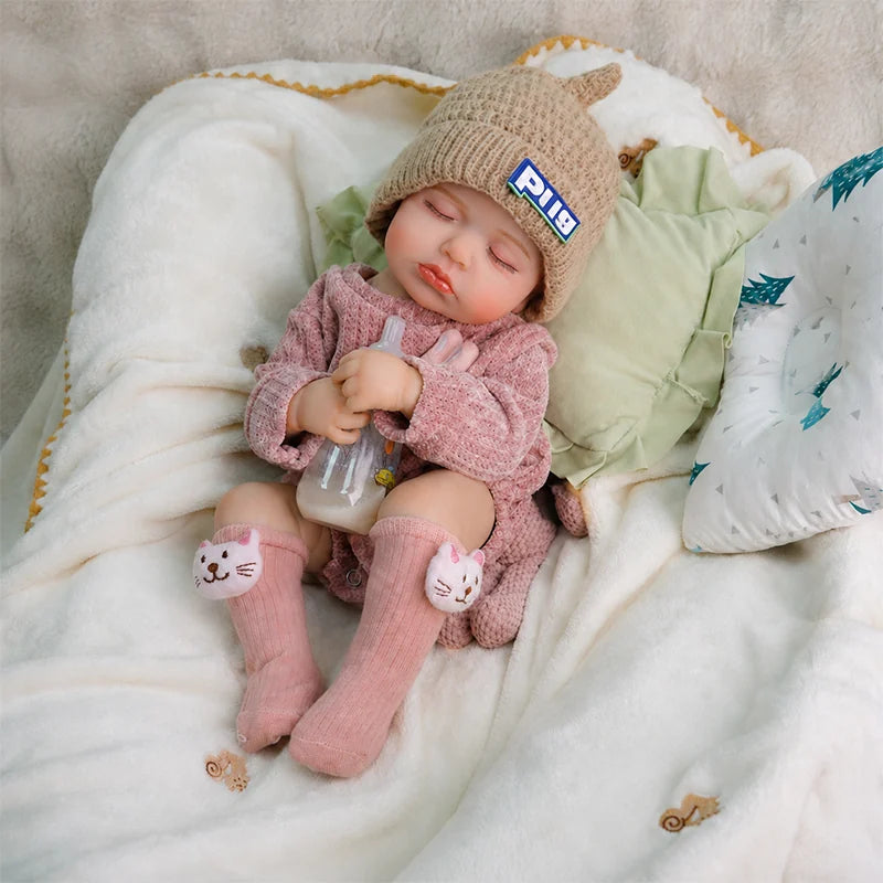 Sleeping reborn baby doll wearing a knitted hat, pink sweater, and cat socks, sitting on a soft blanket while holding a baby bottle.