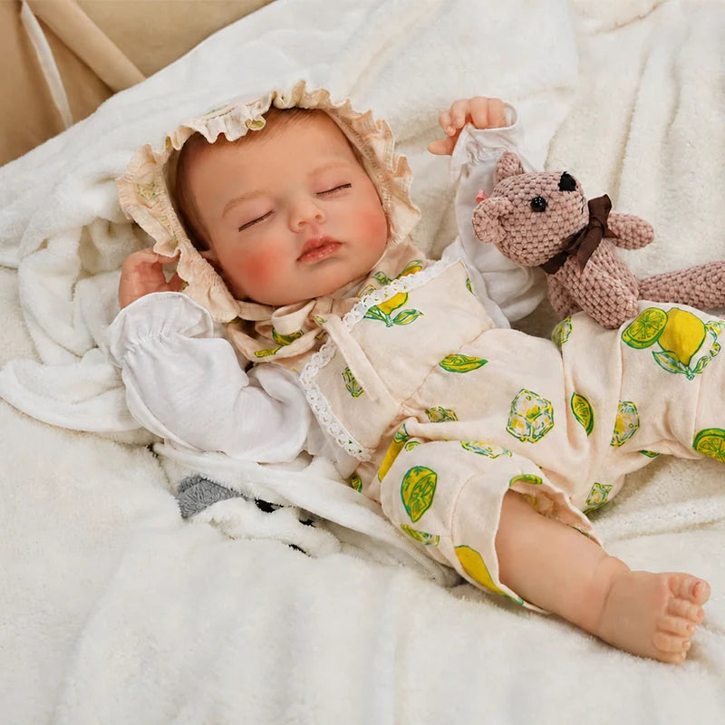 Sleeping reborn baby doll wearing a lemon-print outfit and ruffled bonnet, lying on a soft white blanket with a small knitted teddy bear beside it.
