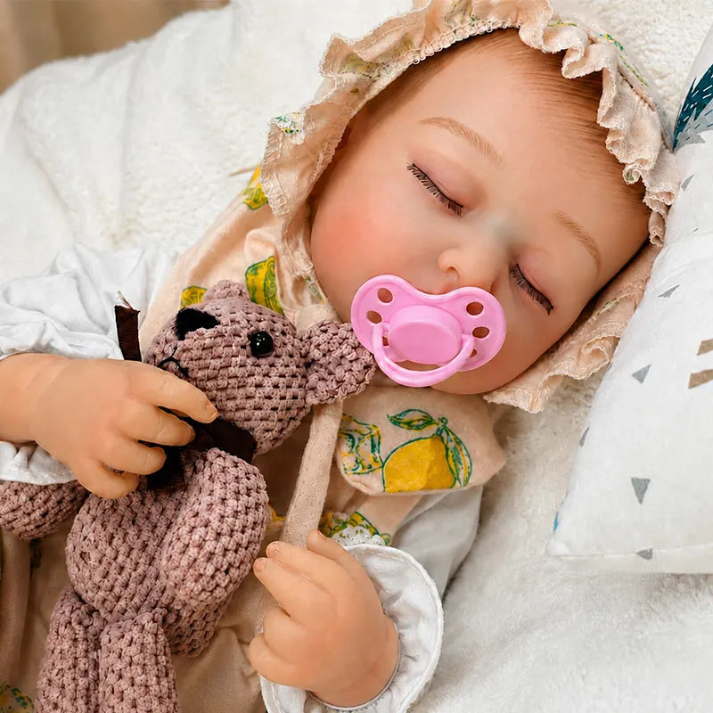 A realistic sleeping reborn baby doll wearing a ruffled lemon-print bonnet rests on a soft white blanket, with a pink pacifier in its mouth and a knitted pink teddy bear toy held against its chest.
