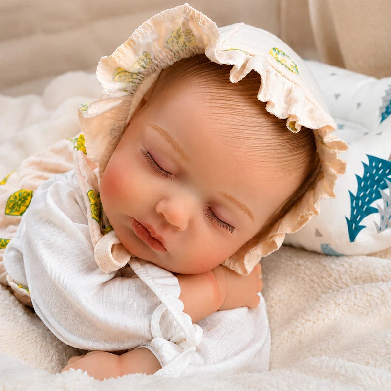 A realistic sleeping reborn baby doll wearing a cream ruffled lemon-print bonnet rests on a soft white blanket, with delicate painted hair, closed eyes, rosy cheeks, and a tree-patterned baby pillow beside it.
