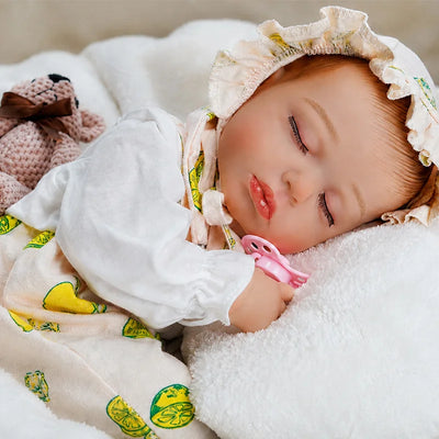 A realistic sleeping reborn baby doll wearing a ruffled lemon-print bonnet and outfit rests on a soft white blanket, holding a pink pacifier with a knitted pink teddy bear beside it.