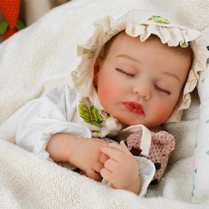 Sleeping reborn baby doll wearing a ruffled bonnet, wrapped in a soft cream blanket and holding a small knitted teddy bear.