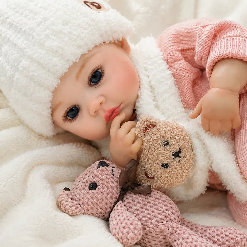 Blue-eyed Rosie reborn baby doll wearing a fluffy white hat and scarf while cuddling a teddy bear and soft plush bunny.