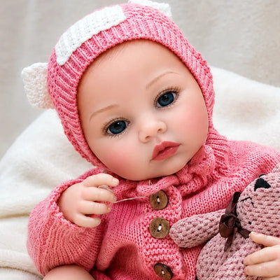 Lifelike reborn baby girl doll wearing a pink knitted romper and matching hat while holding a handmade teddy bear.