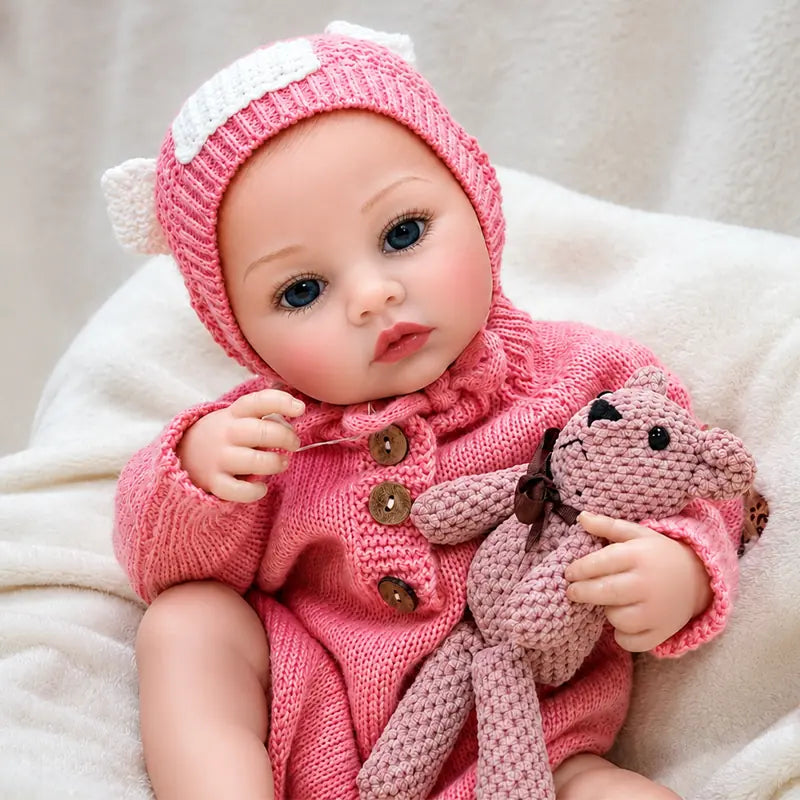 Blue-eyed reborn baby girl in a cozy pink knit outfit, cuddling a small teddy bear with an alert and charming expression.