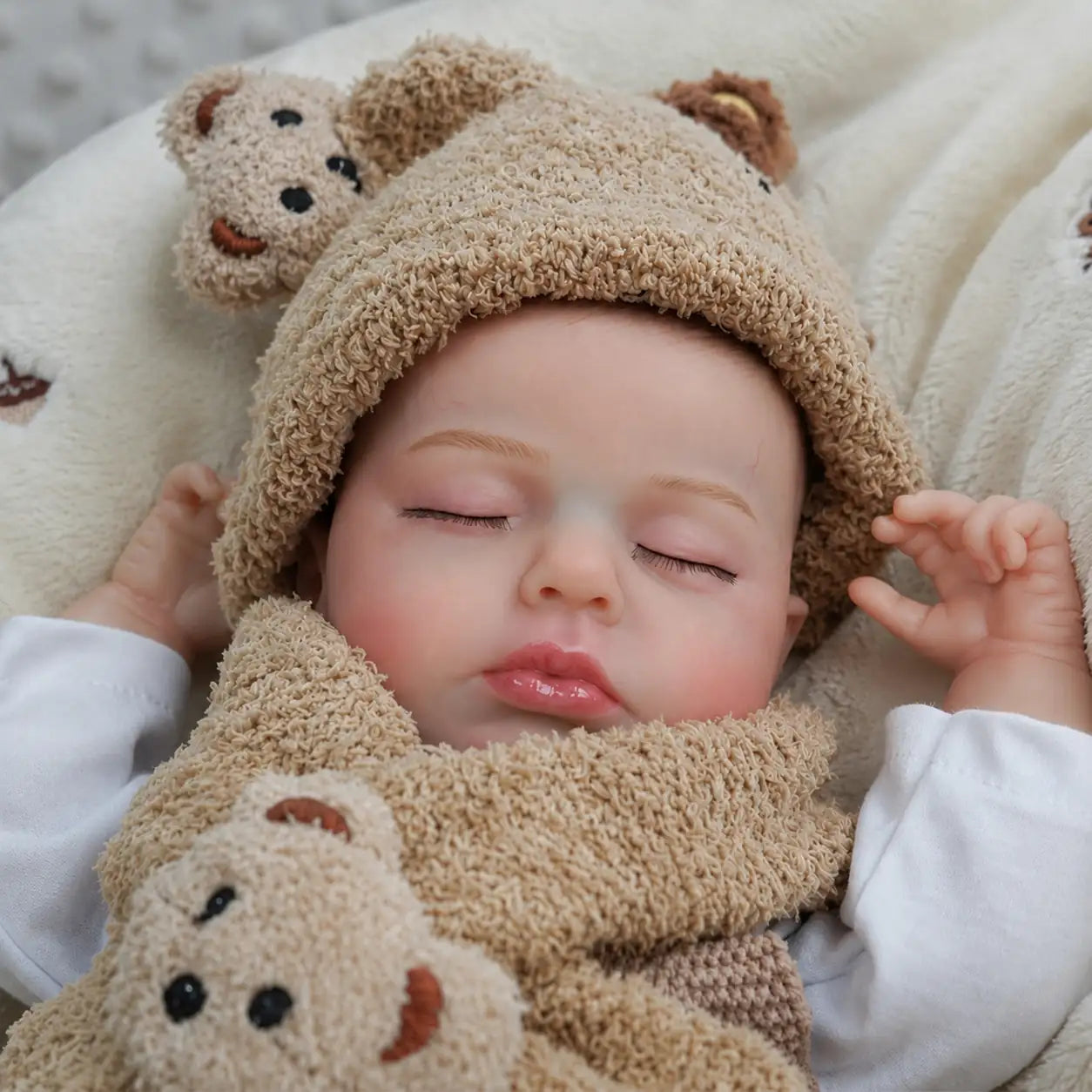A realistic sleeping reborn baby doll wearing a fuzzy teddy bear hood and scarf lies on a soft cream blanket, with closed eyes, rosy cheeks, glossy lips, and a plush bear toy resting against its chest.