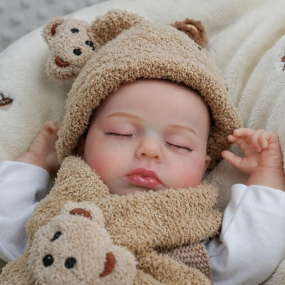 A realistic sleeping reborn baby doll wearing a fuzzy teddy bear hood and scarf lies on a soft cream blanket, with closed eyes, rosy cheeks, glossy lips, and a plush bear toy resting against its chest.
