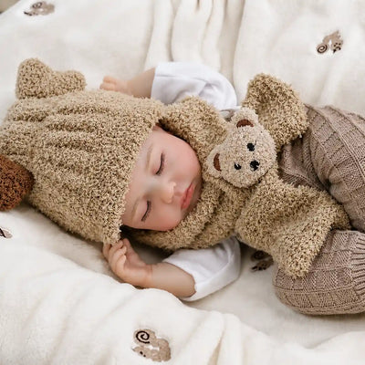A realistic sleeping reborn baby doll dressed in a fuzzy teddy bear hood and scarf lies on a soft cream blanket, wearing taupe knitted pants and cuddling a small plush bear against its chest.