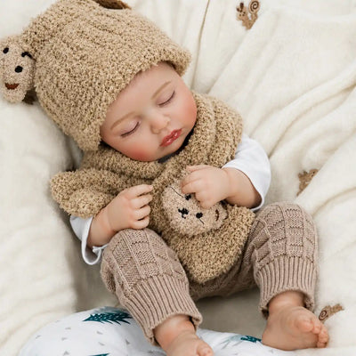 A realistic sleeping reborn baby doll dressed in a fuzzy teddy bear hat and scarf lies on a soft cream blanket, wearing taupe knitted pants and holding a small plush bear on its chest.