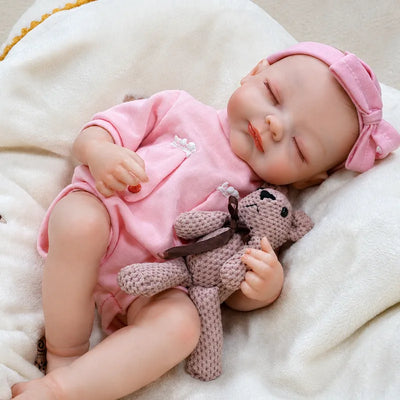Sleeping reborn baby girl in a pink romper and bow headband, gently holding a knitted teddy bear on a cozy blanket.