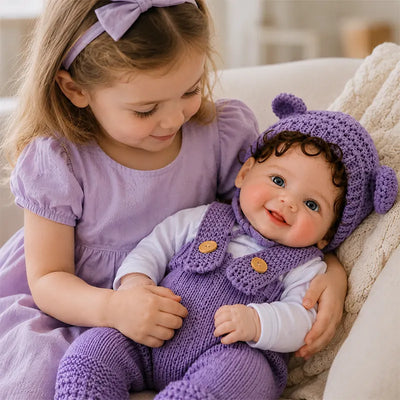 Young girl cuddling a smiling realistic reborn baby doll dressed in a purple knitted romper and matching bear-ear hat.