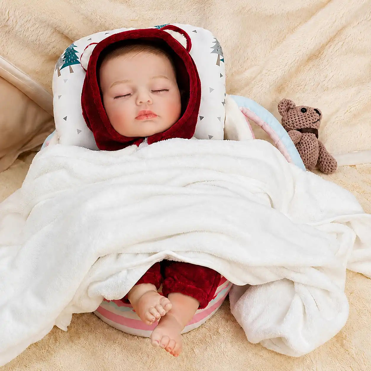 Sleeping reborn baby doll in red hooded outfit, wrapped in white blanket, lying on patterned pillow and beige background.