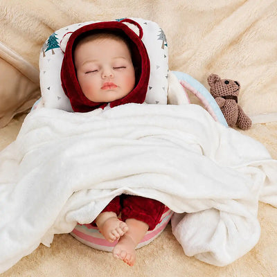 Sleeping reborn baby doll in red hooded outfit, wrapped in white blanket, lying on patterned pillow and beige background.