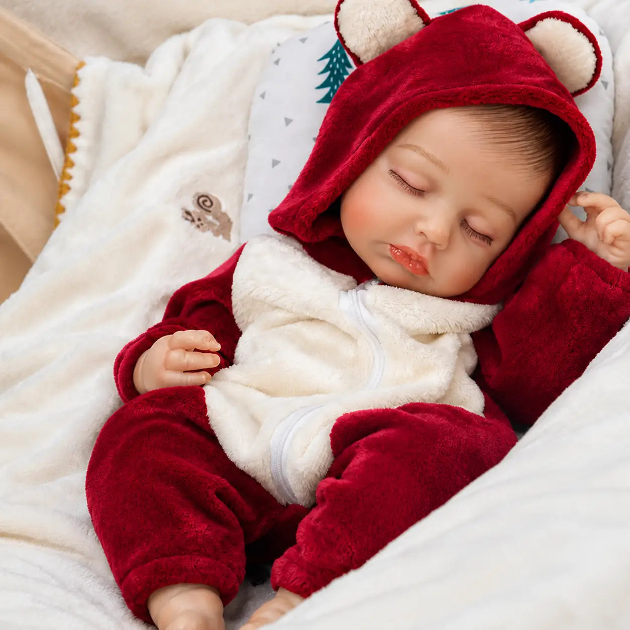 A sleeping baby doll in a red hooded onesie with bear ears, lying on a light blanket.