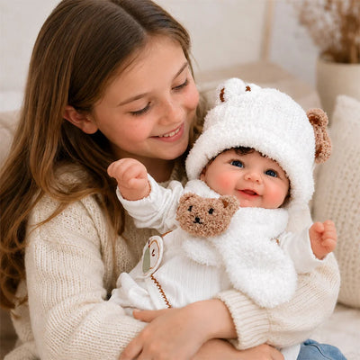 Smiling young girl cuddling a lifelike reborn baby doll dressed in a white teddy bear hat, scarf, and cozy winter outfit.