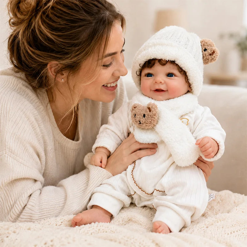 Smiling lifelike reborn baby doll in a cozy white outfit, teddy bear hat, and scarf, lovingly held by a woman in a warm home setting.