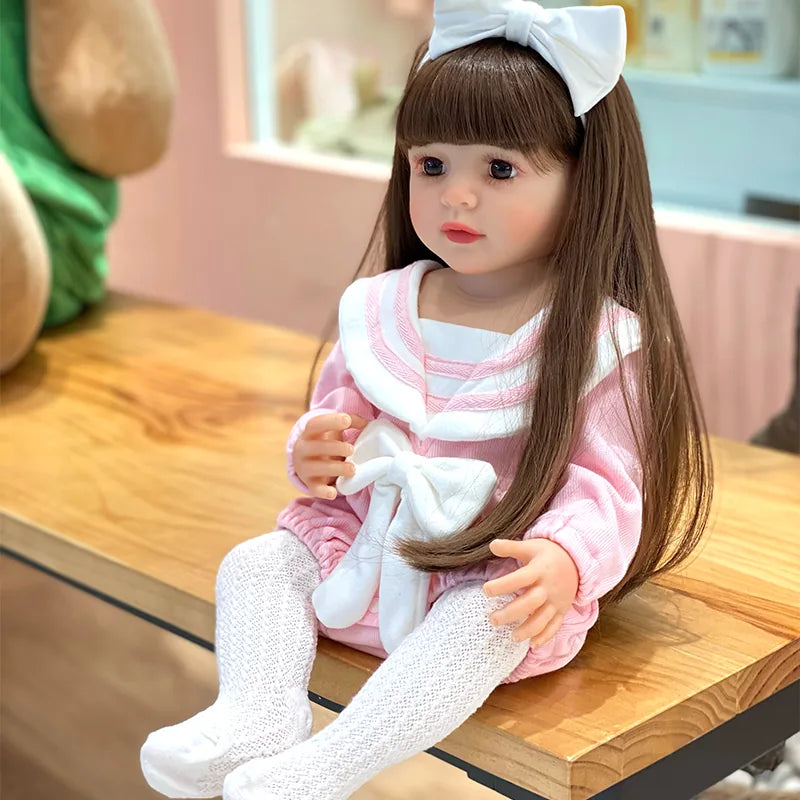 Interactive reborn toddler doll with long brown hair, expressive eyes, a white bow, and a sweet pink outfit.