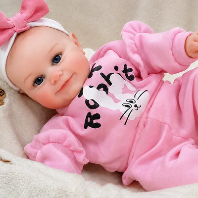 Adorable reborn baby doll relaxing in a cozy nursery setting, wearing a bright pink outfit and bow while smiling sweetly at the camera.