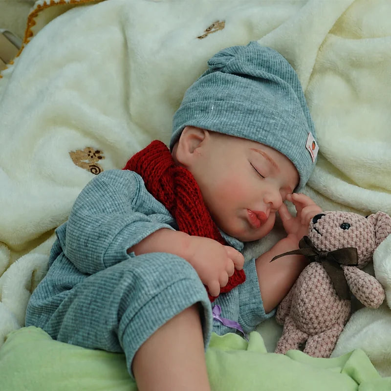 A sleeping reborn baby doll dressed in a blue outfit and matching hat, wearing a red knitted scarf, lying on soft blankets.