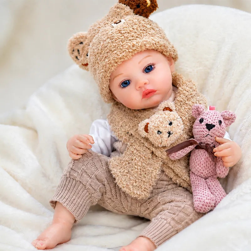 Realistic reborn baby doll cuddling a pink knitted bunny while dressed in a cozy teddy bear winter outfit.