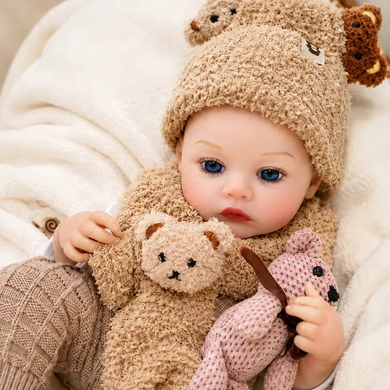 Blue-eyed reborn baby doll wearing a teddy bear hat, matching knitted trousers, and a soft animal-themed scarf.
