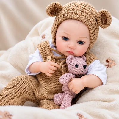 Lifelike reborn baby girl doll with blue eyes, realistic painted features, brown knit clothing, and a pink teddy bear accessory.